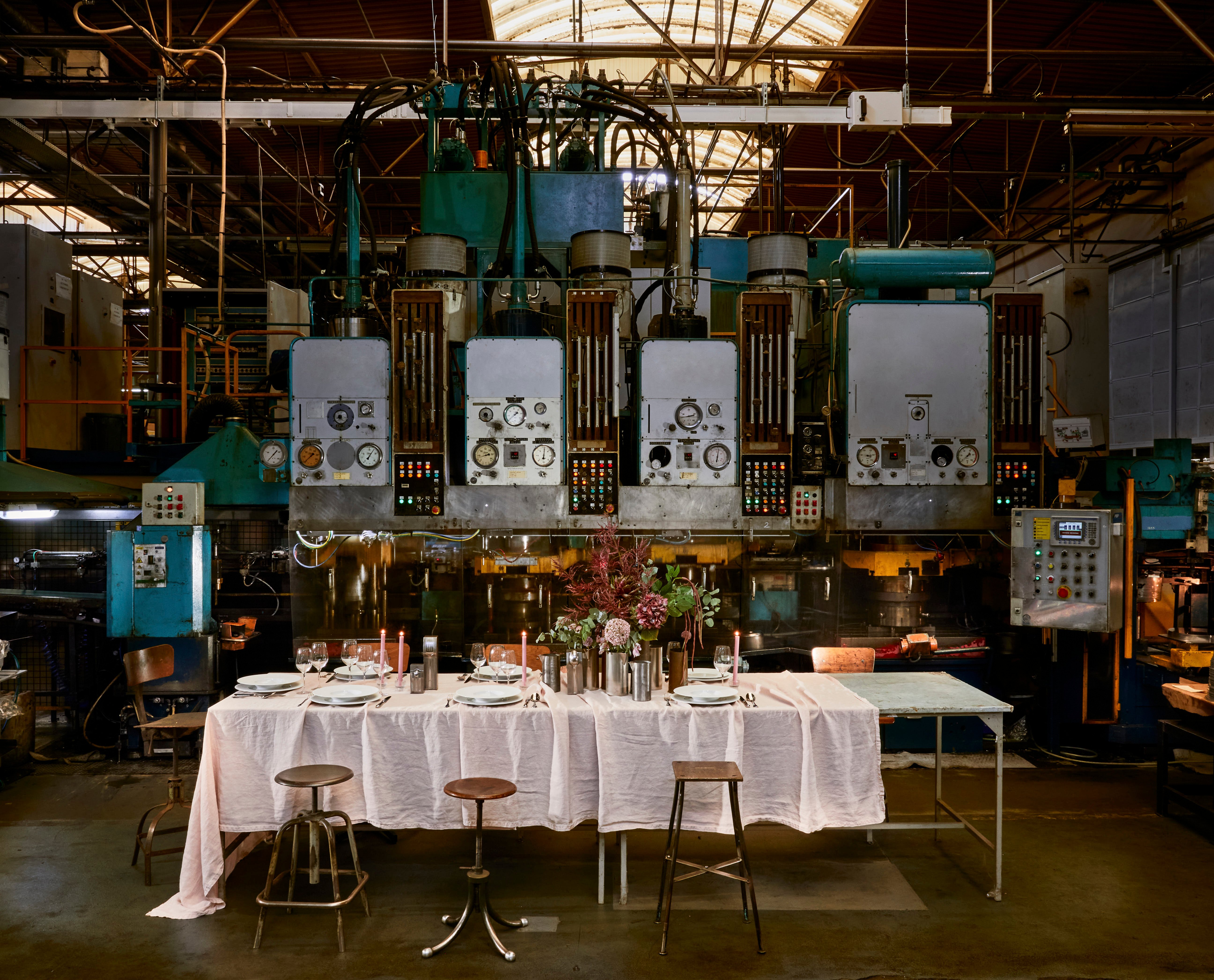 Une table installée devant des machines d'usine avec une nappe blanche, de la vaisselle en porcelaine, des verres à pied et des couverts en inox.